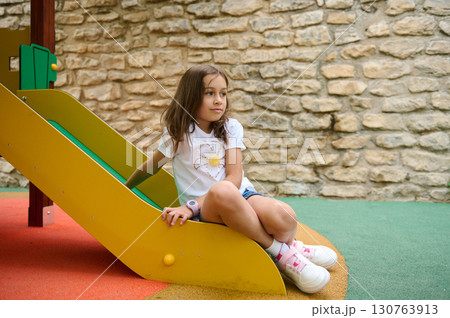 Young Girl Relaxing on Playground Slide Against Stone Wall Background Young Girl Relaxing on Playground Slide Against Stone Wall Background 130763913