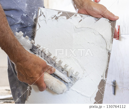 Craftsman spreads tile adhesive with notched trowel on a tile back, closeup. Construction process Craftsman spreads tile adhesive with notched trowel on a tile back, closeup. Construction process 130764112