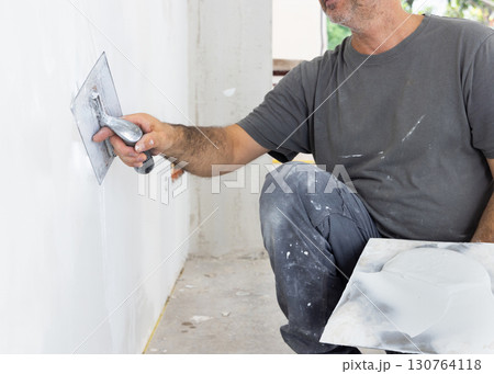 Man in grey work clothes uses trowel and hawk to plaster a wall, construction or renovation of house Man in grey work clothes uses trowel and hawk to plaster a wall, construction or renovation of house 130764118