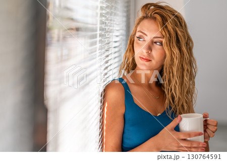 Thoughtful Woman Holding Coffee Cup by Window Blinds 130764591