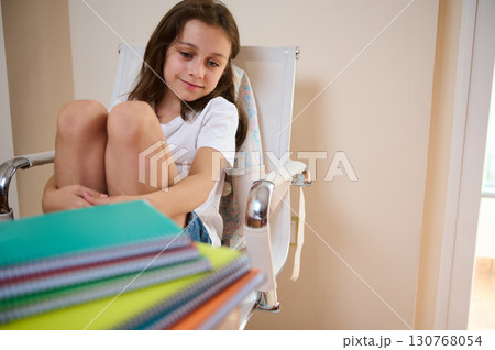 Thoughtful Young Girl Sitting With Colorful Notebooks on a Desk in a Room Thoughtful Young Girl Sitting With Colorful Notebooks on a Desk in a Room 130768054