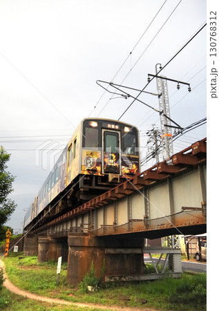 石見神楽のラッピンング列車（キハ126系ディーゼル気動車）が市街地の背の低い橋桁の鉄橋を通過する光景 130768312