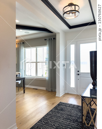 Spacious entryway featuring a coffered ceiling, patterned black rug, and wide windows with curtains. Light walls and natural wood flooring give the space an inviting look. 130768627