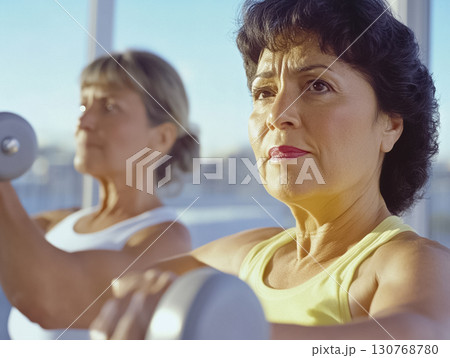 Confident older woman lifting dumbbells in a sunny studio setting. Excellent for promoting healthy aging, women’s fitness, and active living content. 130768780