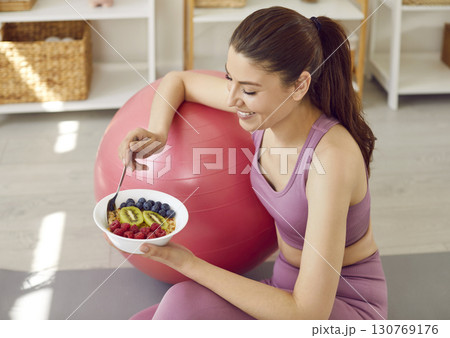 Sporty slim woman in sportswear eating healthy food sitting on yoga mat at home. Sporty slim woman in sportswear eating healthy food sitting on yoga mat at home. 130769176