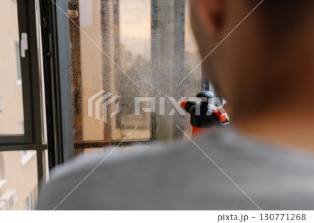 Rear view of janitor male spraying cleaning solution onto large window, preparing to wipe and polish to sparkling shine, ensuring clear and unobstructed view of city outside. Concept of domestic work. 130771268