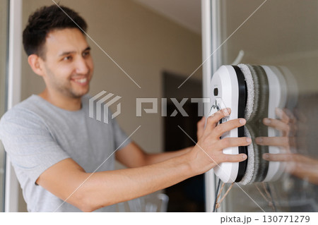 Selective focus shot of smiling young man installing robotic vacuum cleaner on window, preparing to activate device for efficient cleaning session, ensuring spotless and sparkling interior environment Selective focus shot of smiling young man installing robotic vacuum cleaner on window, preparing to activate device for efficient cleaning session, ensuring spotless and sparkling interior environment 130771279