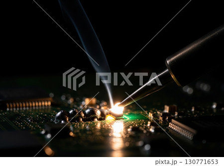 Soldering iron tip melting solder on a green pcb with smoke in a dark electronics workshop 130771653