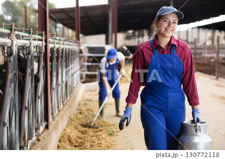 Young positive female farmer carrying milk churn near stall with cows during work on dairy farm Young positive female farmer carrying milk churn near stall with cows during work on dairy farm 130772118