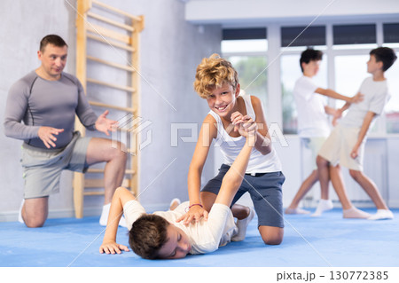 Concentrated boy learning effective self defence techniques in sparring, practicing painful pronating wristlock on male opponent lying face down on floor at sports studio 130772385