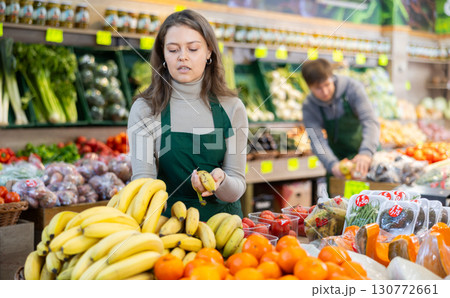 Young female seller holding bananas standing by counter in grocery market 130772661