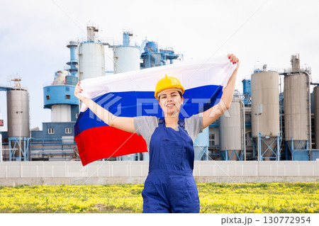 Young female engineer in helmet waving state flag of Russia while standing in front of big tanks at chemical plant Young female engineer in helmet waving state flag of Russia while standing in front of big tanks at chemical plant 130772954