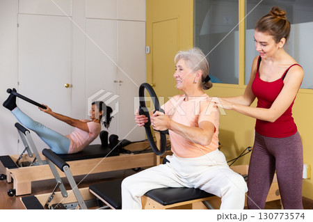 Young trainer helping elderly woman exercising with resistance ring Young trainer helping elderly woman exercising with resistance ring 130773574