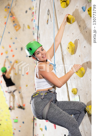 Sporty young girl in safety gear doing difficult wall climb in climbing gym 130773799