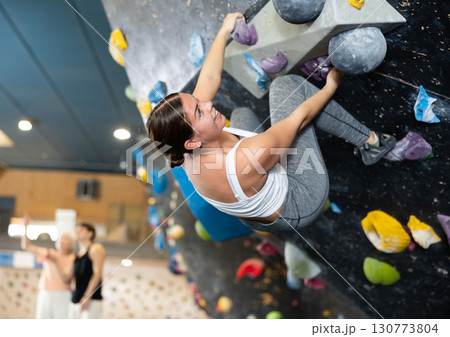 Confident young woman scaling artificial climbing wall in climbing gym 130773804