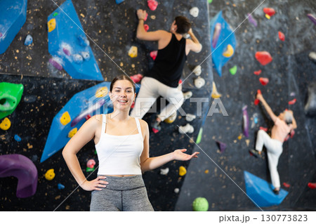 Fit young woman in sportswear smiling at camera while standing against artificial training wall in adventure park or rock climbing gym 130773823