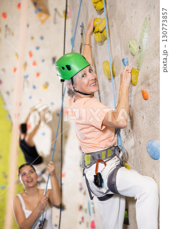 Determined older woman in safety helmet doing her best at climbing artificial wall Determined older woman in safety helmet doing her best at climbing artificial wall 130773857