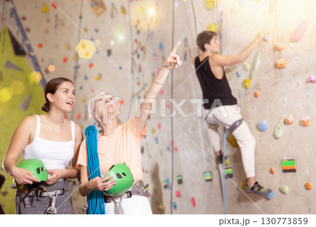 Two women of different ages standing with blue safety belts and helmet before practicing rock-climbing on climbing wall in adventure park Two women of different ages standing with blue safety belts and helmet before practicing rock-climbing on climbing wall in adventure park 130773859