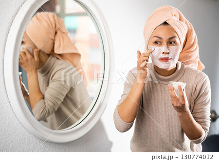 Woman looking at mirror and smoothing cream over face 130774012
