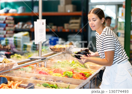 Smiling young female shopper choosing ripe eggplants in garden produce store 130774493