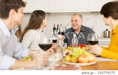 Senior man at kitchen table with wife, grown son and daughter, enjoying conversation at festive table with wine and snacks Senior man at kitchen table with wife, grown son and daughter, enjoying conversation at festive table with wine and snacks 130774575