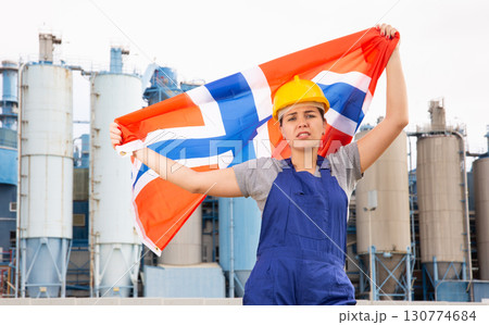 Upset girl in overalls with Norway flag in her hands against the background of chimneys of modern factory 130774684