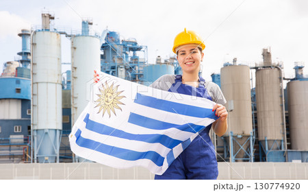 Young female engineer in helmet waving state flag of Uruguay while standing in front of big tanks at chemical plant Young female engineer in helmet waving state flag of Uruguay while standing in front of big tanks at chemical plant 130774920
