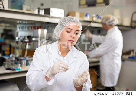 Professional female chemist in uniform examining solution in petri dish while working in research center Professional female chemist in uniform examining solution in petri dish while working in research center 130774921