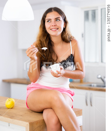 Young woman sitting on kitchen table and eating yoghurt Young woman sitting on kitchen table and eating yoghurt 130774956