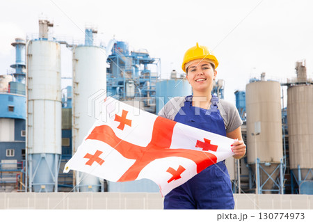 National flag of Georgia in the hands of girl in overalls against background of modern metallurgical plant National flag of Georgia in the hands of girl in overalls against background of modern metallurgical plant 130774973