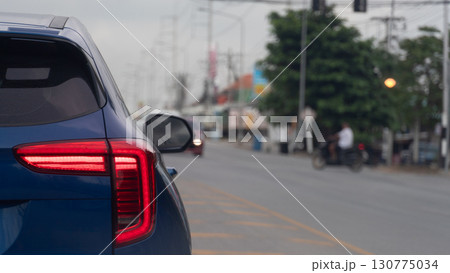 Rear side of blue car with turn on brake light. Traffic in the junction of asphalt road. Blurred of city with motorcycle and driver and trees with city under soft sky. 130775034