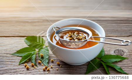 Close-up of hot herbal tea in a white bowl. A strainer with fresh stevia leaves is visible in the drink. This photo, taken in natural soft light on an aged wooden table, highlights the natural beauty  Close-up of hot herbal tea in a white bowl. A strainer with fresh stevia leaves is visible in the drink. This photo, taken in natural soft light on an aged wooden table, highlights the natural beauty  130775688