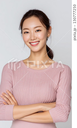 Smiling Korean woman in a light pink sweater poses confidently with arms crossed against a neutral background Smiling Korean woman in a light pink sweater poses confidently with arms crossed against a neutral background 130777250