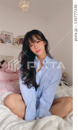 Korean woman in a blue striped shirt sitting on a bed surrounded by soft pillows in a cozy room Korean woman in a blue striped shirt sitting on a bed surrounded by soft pillows in a cozy room 130777286