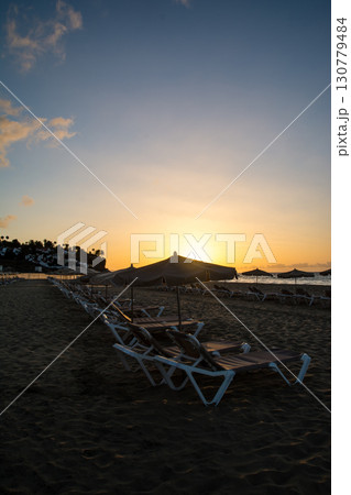 水平線から昇るまぶしい朝日と海岸線の砂浜に並べられたビーチベッドとパラソル　フエルテベンテュラ島にて 130779484
