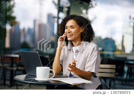 A Woman Engaged in Working on a Laptop while Taking a Phone Call in a Cozy Cafe Setting A Woman Engaged in Working on a Laptop while Taking a Phone Call in a Cozy Cafe Setting 130780055