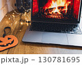 Notebook with flame on screen with garland and decorative pumpkin on wooden table. Halloween still life. Cozy lifestyle concept. Autumn lifestyle. Bonfire on notebook and garland. October background.  130781695