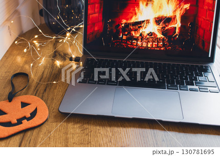 Notebook with flame on screen with garland and decorative pumpkin on wooden table. Halloween still life. Cozy lifestyle concept. Autumn lifestyle. Bonfire on notebook and garland. October background.  130781695