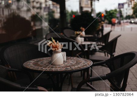 A charming cafe terrace with tables, chairs, and a vase of dried flowers adds a touch of charm awaits customers. An inviting outdoor cafe scene. A charming cafe terrace with tables, chairs, and a vase of dried flowers adds a touch of charm awaits customers. An inviting outdoor cafe scene. 130782064
