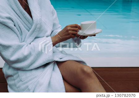 A woman, girl in a white bathrobe holding a cup, enjoying a moment of relaxation. A person in a relaxed setting by a swimming pool in modern a hotel. A woman, girl in a white bathrobe holding a cup, enjoying a moment of relaxation. A person in a relaxed setting by a swimming pool in modern a hotel. 130782070