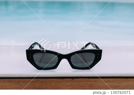Elegant funky black sunglasses displayed by a pool, capturing tranquil summer scene. Stylish sunglasses rest on a white surface against blurred water. Elegant funky black sunglasses displayed by a pool, capturing tranquil summer scene. Stylish sunglasses rest on a white surface against blurred water. 130782071