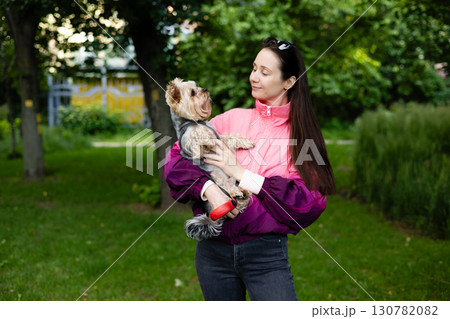 A woman embraces her adorable yorkshire terrier in a park setting, sharing a loving moment. A woman gazes lovingly at her Yorkshire Terrier as she holds the dog in her arms outdoors in a park. 130782082