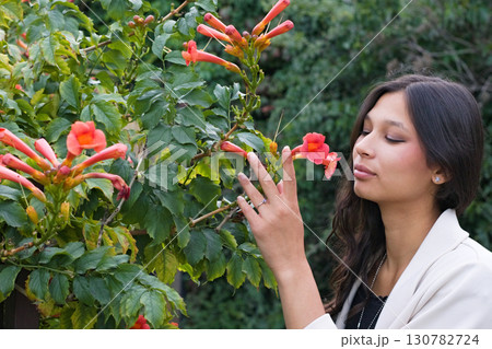 Young Indian woman with long dark hair standing near orange blossoms, gently raising her hand toward the flowers. A lifestyle and cultural beauty portrait 130782724
