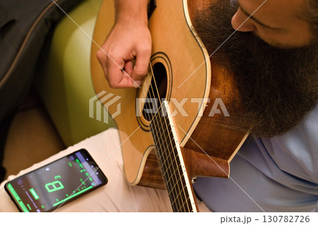 Bearded man tuning acoustic guitar using smartphone tuner app, close up of hands and instrument, modern combination of traditional music practice and digital technology. 130782726