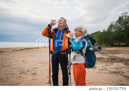 Hiking tourism adventure. Senior couple man woman drinking water enjoying outdoor recreation hiking on beach. Old people backpackers hikers enjoy walking hike trekking active vacation beauty of nature Hiking tourism adventure. Senior couple man woman drinking water enjoying outdoor recreation hiking on beach. Old people backpackers hikers enjoy walking hike trekking active vacation beauty of nature 130782739