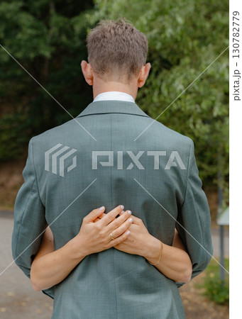 A couple shares a quiet embrace, the womans arms wrapped around the man's back. They are outdoors, surrounded by greenery, wearing formal attire, conveying intimacy and connection A couple shares a quiet embrace, the womans arms wrapped around the man's back. They are outdoors, surrounded by greenery, wearing formal attire, conveying intimacy and connection 130782779