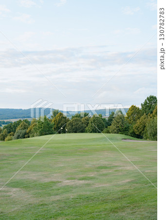 Expansive golf course with vibrant green grass, surrounded by lush trees under a partly cloudy sky. Two flags indicate the holes, creating a serene and tranquil setting 130782783