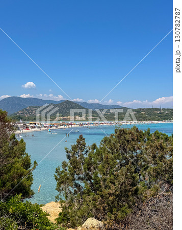 Sunny summer day at a popular beach in Sardinia with turquoise waters, sunbathers, and distant green mountains. Sunny summer day at a popular beach in Sardinia with turquoise waters, sunbathers, and distant green mountains. 130782787