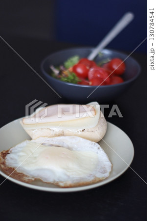 breakfast scene: fried egg and open sandwich on a beige plate, with a blurred bowl of cherry-tomato salad in the background. Soft daylight and shallow depth of field create a cozy, home-cooking mood. 130784541