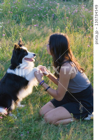 Woman kneeling on grass holding the paws of a Border Collie dog during training in a sunny field. Ideal for pet training, obedience lessons, companionship, and outdoor lifestyle themes. Woman kneeling on grass holding the paws of a Border Collie dog during training in a sunny field. Ideal for pet training, obedience lessons, companionship, and outdoor lifestyle themes. 130784549